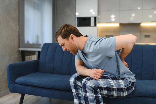 Photo people healthcare and treatment portrait of sick man suffering from acute side back pain sitting on couch at home person touching lower back aching area with hand health problem concept