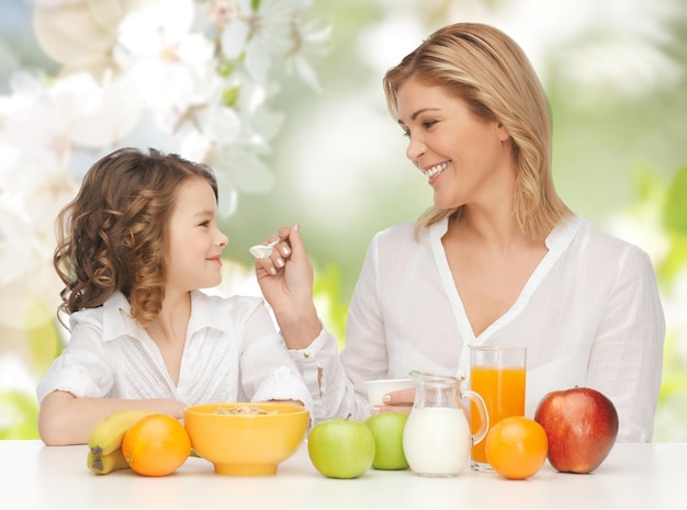 people, healthy lifestyle, family and food concept - happy mother and daughter eating healthy breakfast over green summer garden background