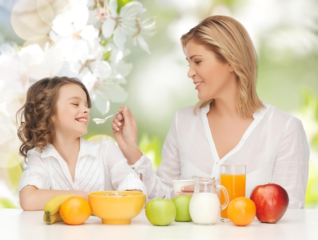 People, healthy lifestyle, family and food concept - happy mother and daughter eating healthy breakfast over green summer garden background