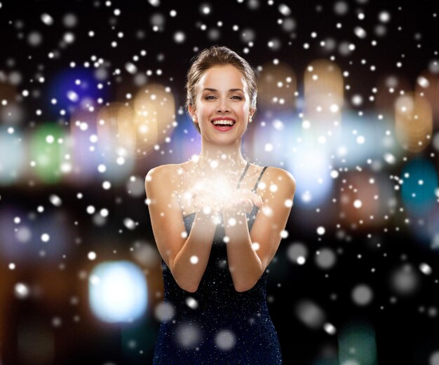 people, holidays, advertisement, christmas and luxury concept - laughing woman in evening dress holding something imaginary over night lights and snow background