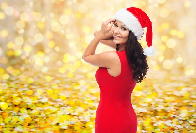 People, holidays, christmas and celebration concept - beautiful sexy woman in santa hat and red dress over yellow lights or golden confetti background