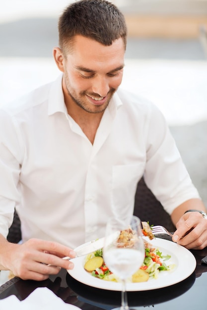 people, holidays, food and leisure concept - happy man with fork and knife eating salad for dinner at restaurant terrace