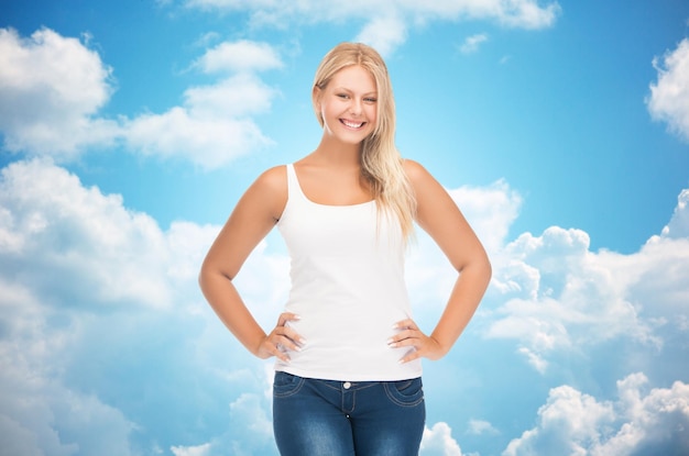 people, holidays, style and body type concept - smiling young woman in blank white shirt and jeans over blue sky and clouds background