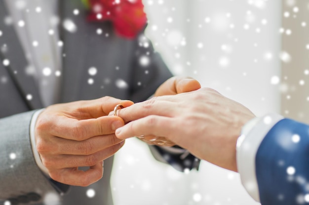 people, homosexuality, same-sex marriage and love concept - close up of happy male gay couple hands putting wedding ring on over snow effect