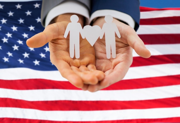 people, homosexuality, same-sex marriage and love concept - close up of happy male gay couple holding paper cutout love symbol over american flag background