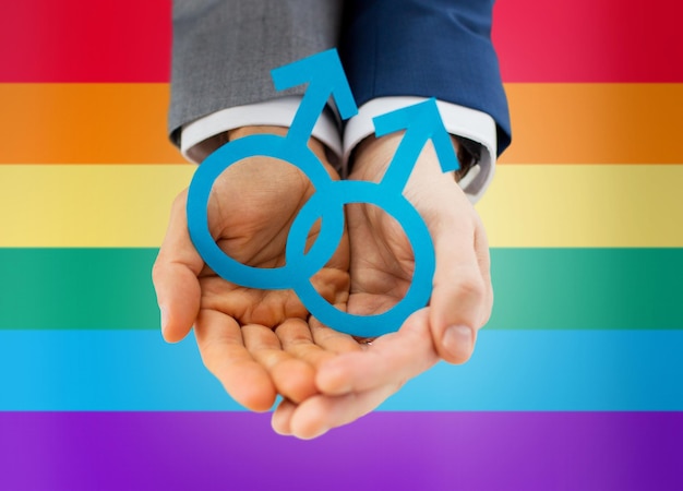 people, homosexuality, same-sex marriage and love concept - close up of happy male gay couple holding paper cutout love symbol over rainbow flag background