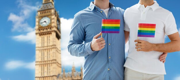 People, Homosexuality, Same-sex Marriage, Travel And Love Concept - Close Up Of Happy Male Gay Couple Holding Rainbow Flags And Hugging From Back Over Big Ben Tower In London Background