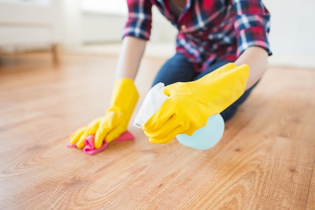 people, housework and housekeeping concept - close up of woman in rubber glover with cloth and detergent spray cleaning floor at home