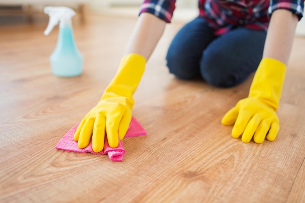 people, housework and housekeeping concept - close up of woman in rubber glover with cloth and detergent spray cleaning floor at home