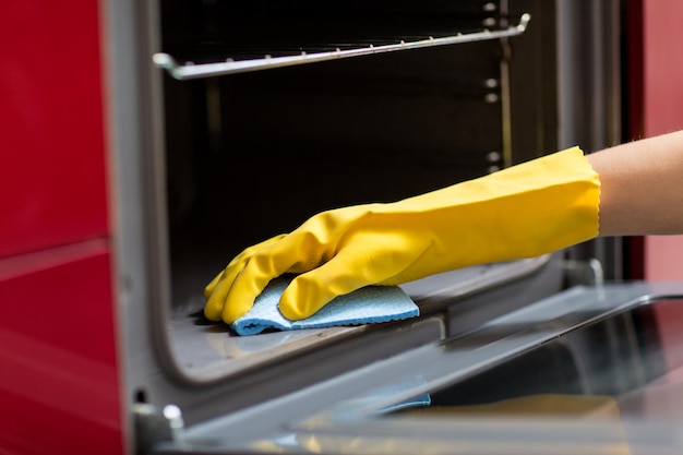 people, housework and housekeeping concept - hand in rubber glove with rag cleaning oven at home kitchen