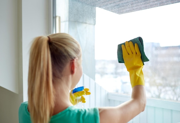 people, housework and housekeeping concept - happy woman in gloves cleaning window with rag and cleanser spray at home