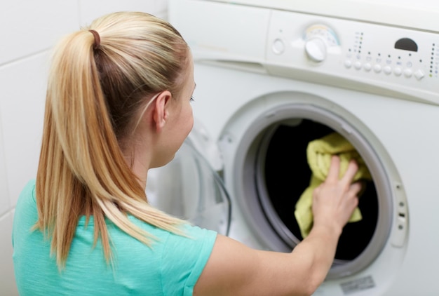 people, housework, laundry and housekeeping concept - happy woman putting laundry into washing machine at home