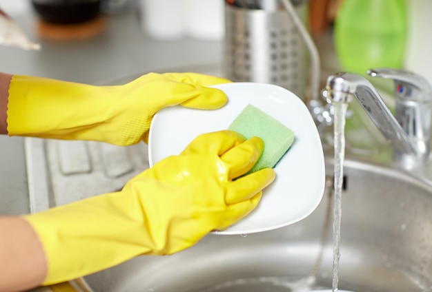 people, housework, washing-up and housekeeping concept - close up of woman hands in protective gloves washing dishes with sponge at home kitchen