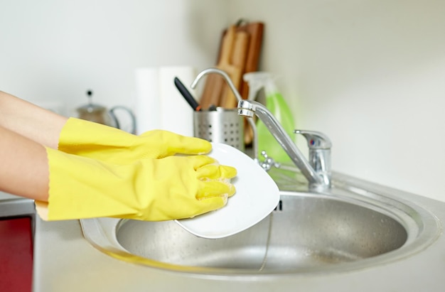 people, housework, washing-up and housekeeping concept - close up of woman hands in protective gloves washing dishes with sponge at home kitchen