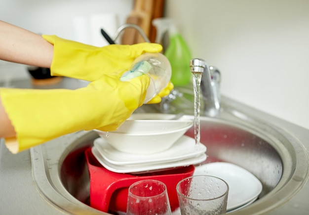 people, housework, washing-up and housekeeping concept - close up of woman hands in protective gloves washing dishes with sponge at home kitchen