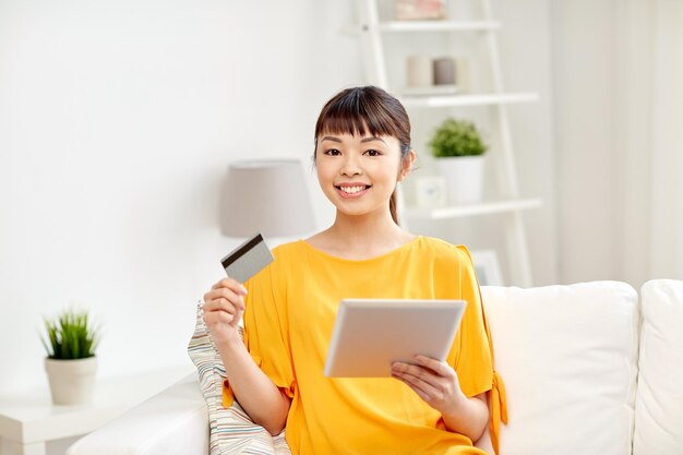 people, internet bank, online shopping, technology and e-money concept - happy asian young woman sitting on sofa with tablet pc computer and credit card at home