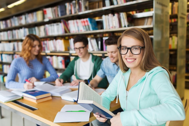 people, knowledge, education, literature and school concept - happy young woman in eyeglasses reading book and preparing to exams over group of students in library