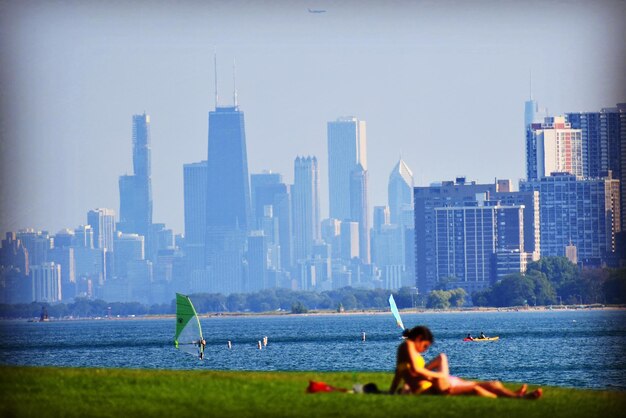 People on lake by modern buildings against sky in summer