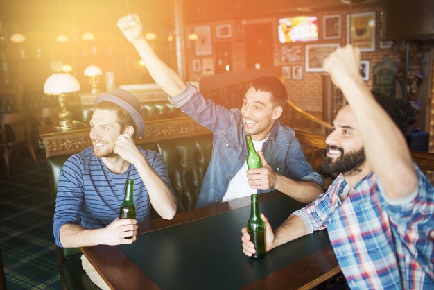 people, leisure, friendship and bachelor party concept - happy male friends drinking bottled beer and raised hands rooting for football match at bar or pub