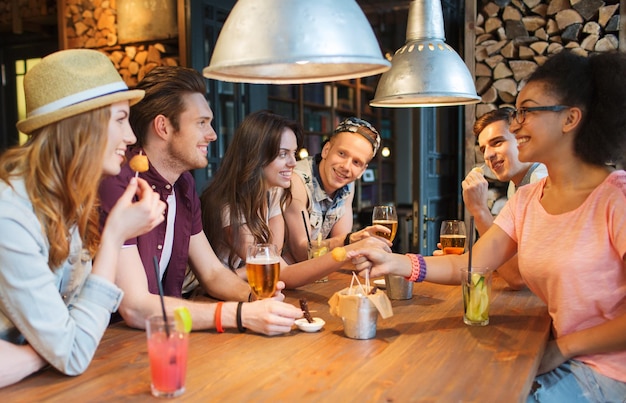 people, leisure, friendship and communication concept - group of happy smiling friends drinking beer and cocktails eating and talking at bar or pub