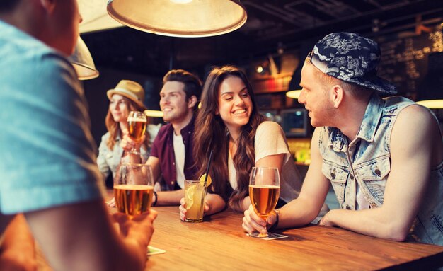 people, leisure, friendship and communication concept - group of happy smiling friends drinking beer and cocktails talking at bar or pub