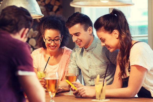 people, leisure, friendship, technology and communication concept - group of happy smiling friends with smartphones and drinks at bar or pub