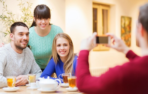 人々、レジャー、友情、テクノロジーのコンセプト-スマートフォンで写真を撮ったり、カフェでお茶を飲んだりする幸せな友達のグループ