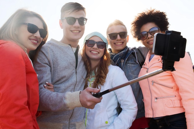 People, leisure, friendship and technology concept - group of\
smiling teenage friends taking picture with smartphone on selfie\
stick outdoors