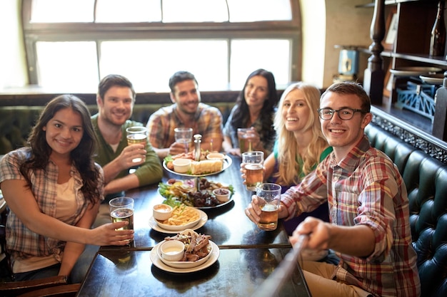 people, leisure, friendship and technology concept - happy friends taking picture by selfie stick, drinking beer and eating snacks at bar or pub