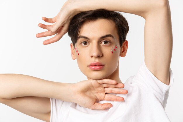 People, lgbtq and beauty concept. Close-up of beautiful androgynous male model with glitter on face, posing sensual at camera, standing over white.