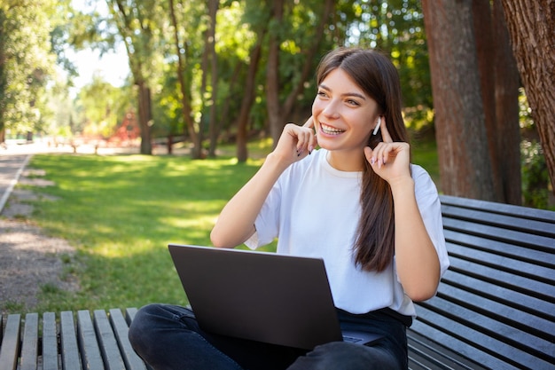 People lifestyle education and technology concept Young happy student girl sitting on bench in park with laptop in wireless headphones enjoys favorite song from playlist Mock up copy space