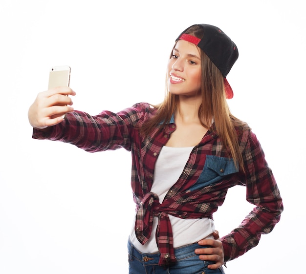 People, lifestyle and tehnology concept: pretty teen girl wearing hat, taking selfies with her smart phone - isolated on white