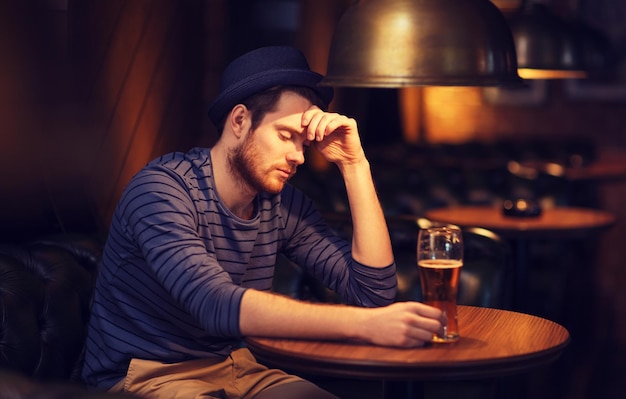 人、孤独、アルコール、ライフスタイルのコンセプト – バーやパブでビールを飲みながら帽子をかぶった不幸な独身青年
