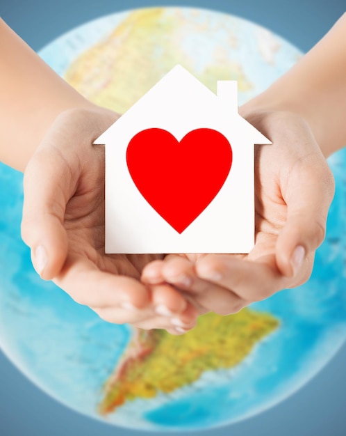 people, love, health, environment and charity concept - close up of human hands holding paper house with red heart over earth globe and blue background