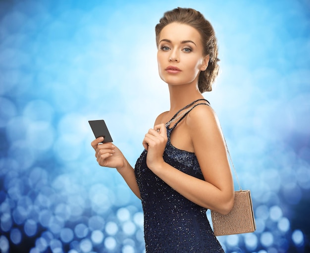 People, luxury, night life and finance concept - beautiful woman in evening dress with vip card and bag over blue lights background