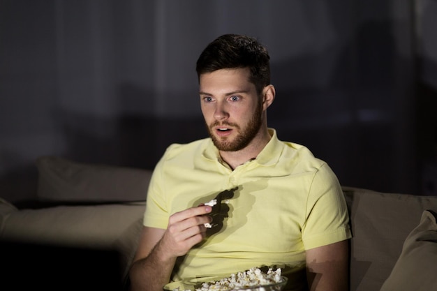 people, mass media, television and entertainment concept - young man watching tv and eating popcorn at night at home