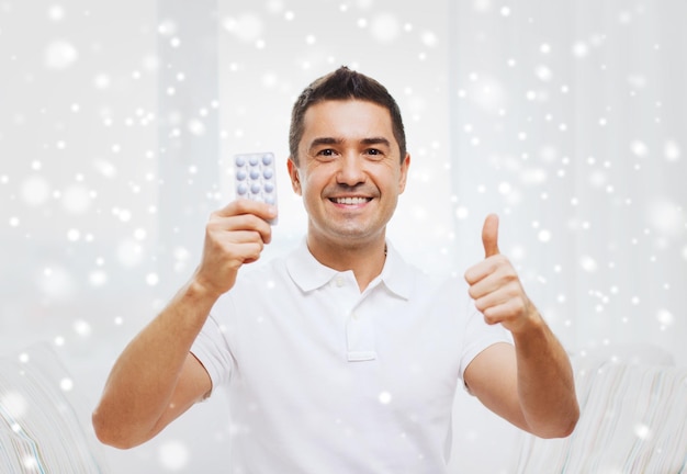people, medication, medicine and health care concept - happy man showing thumbs up and holding pack of pills at home over snow effect