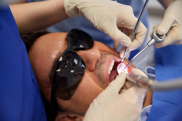 People, medicine, stomatology and health care concept - close\
up of dentist and assistant hands with dental mirror, drill and air\
water gun spray treating male patient teeth at dental clinic