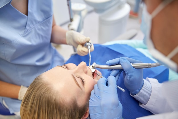 People, medicine, stomatology and health care concept - close\
up of dentists and assistant with mirror, drill and dental air\
water gun spray treating female patient teeth at dental clinic