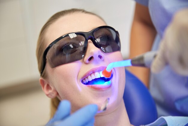 People, medicine, stomatology and health care concept - close\
up of woman patient in protective eyeglasses or goggles with dental\
curing light treating teeth at dental clinic office