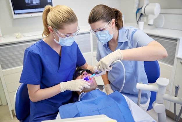 Concetto di persone, medicina, stomatologia e assistenza sanitaria - dentista e assistente femminile con lampada per polimerizzazione dentale e specchio per il trattamento dei denti del paziente maschile presso l'ufficio della clinica dentale