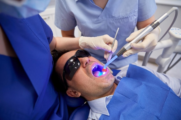 people, medicine, stomatology and health care concept - female dentist and assistant with dental curing light and mirror treating male patient teeth at dental clinic office