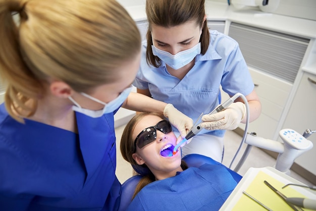 People, medicine, stomatology and health care concept - female\
dentists with dental curing light and mirror treating patient girl\
teeth at dental clinic office