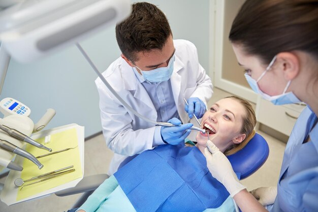 人、医学、口腔病学、ヘルスケアのコンセプト – 男性の歯科医とデンタルミラー、ドリル、エアウォーターガンスプレーを使用した助手が歯科医院で女性患者の歯を治療