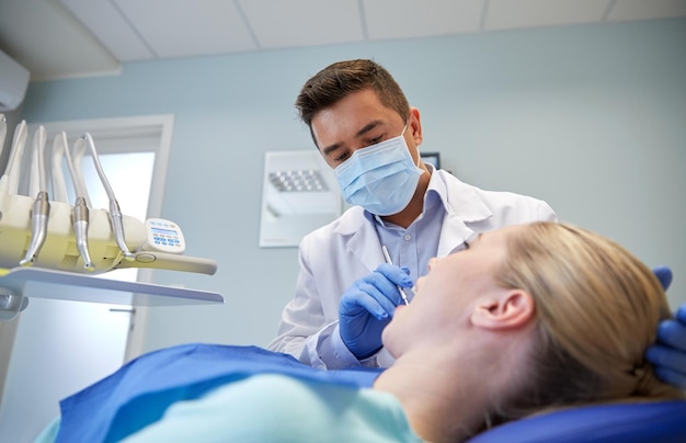 people, medicine, stomatology and health care concept - male dentist in mask checking female patient teeth up at dental clinic office