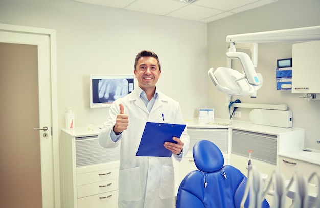 people, medicine, stomatology and healthcare concept - happy middle aged male dentist in white coat with clipboard showing thumbs up at dental clinic office