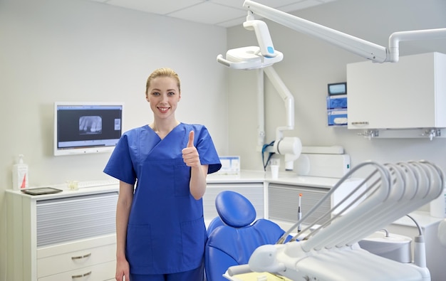 people, medicine, stomatology and healthcare concept - happy young female dentist showing thumbs up at dental clinic office