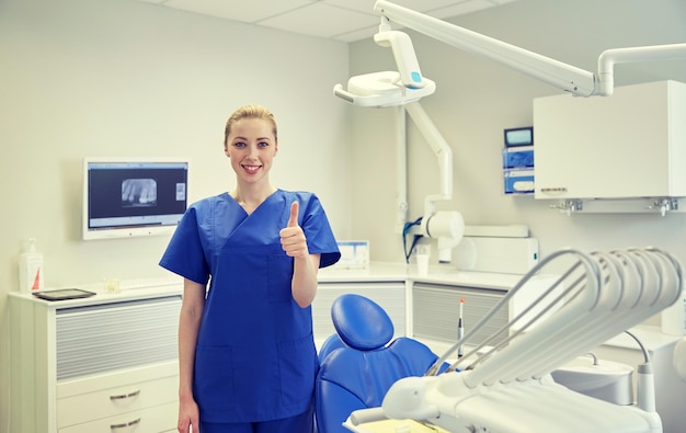 people, medicine, stomatology and healthcare concept - happy young female dentist showing thumbs up at dental clinic office