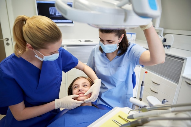 people, medicine, stomatology and teeth care concept - happy female dentist with assistant checking patient girl dental occlusion teeth at dental clinic office