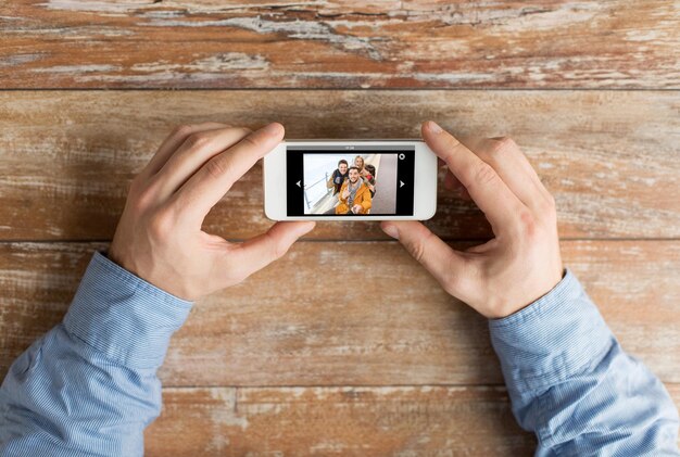 people, memory, relations and technology concept - close up of male hands holding smartphone with photo of happy friends on screen at table